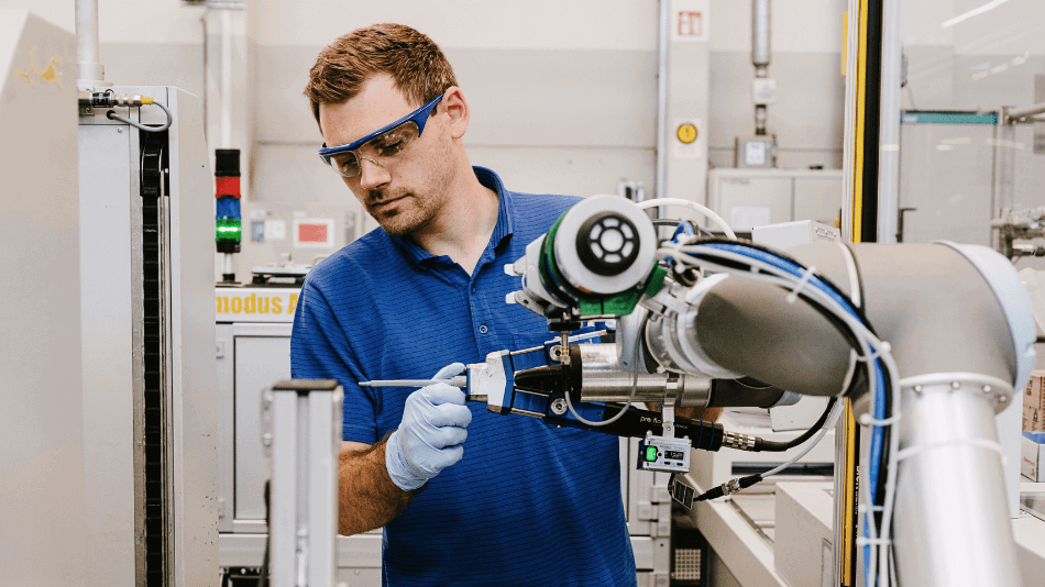 Ein Roboterarm steht im Vordergrund mit einer Klebepistole ausgestattet. Hinter ihm platziert ein Mitarbeiter in einem blauen T-Shirt die Spitze der Klebepistole präzise.